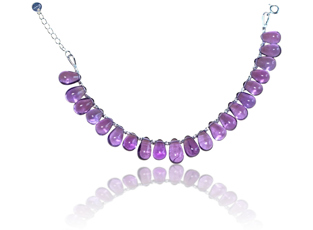 The Fire bracelet laid open in a curve on a dark surface: purple teardrop stones and their reflection, with the spring-ring clasp, extender chain and round River E tag